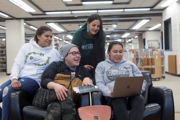 A group of four Indigenous students in the ITEP program are seated or standing behind a chair in the library reviewing information on a laptop computer